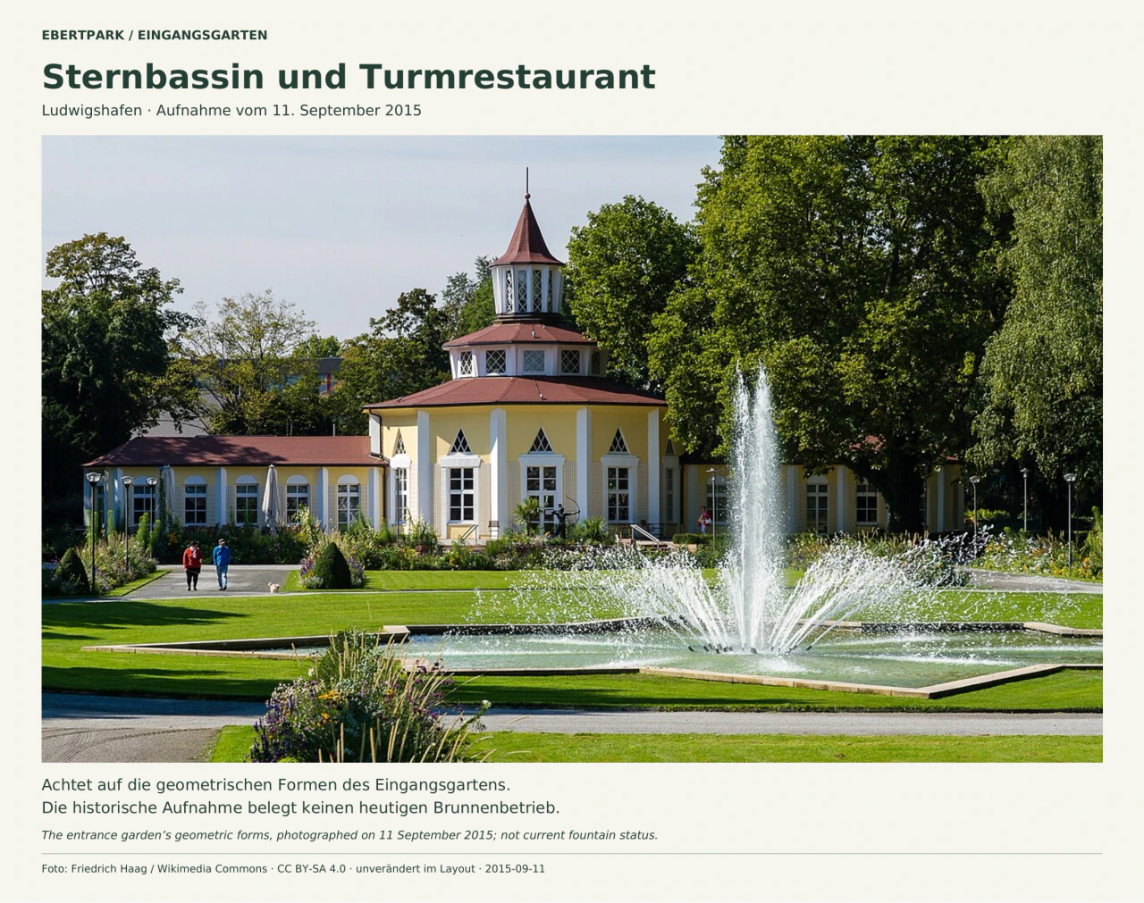 Ein sternförmiges flaches Wasserbecken mit einer hohen Fontäne und seitlich ausstrahlenden Wasserstrahlen liegt vor dem gelben Turmrestaurant. Das Gebäude hat ein gestuftes rotes Dach und einen schlanken Turm; grüne Rasenflächen, Staudenbeete und große Laubbäume umgeben den Platz. Einige Menschen gehen auf den Wegen.