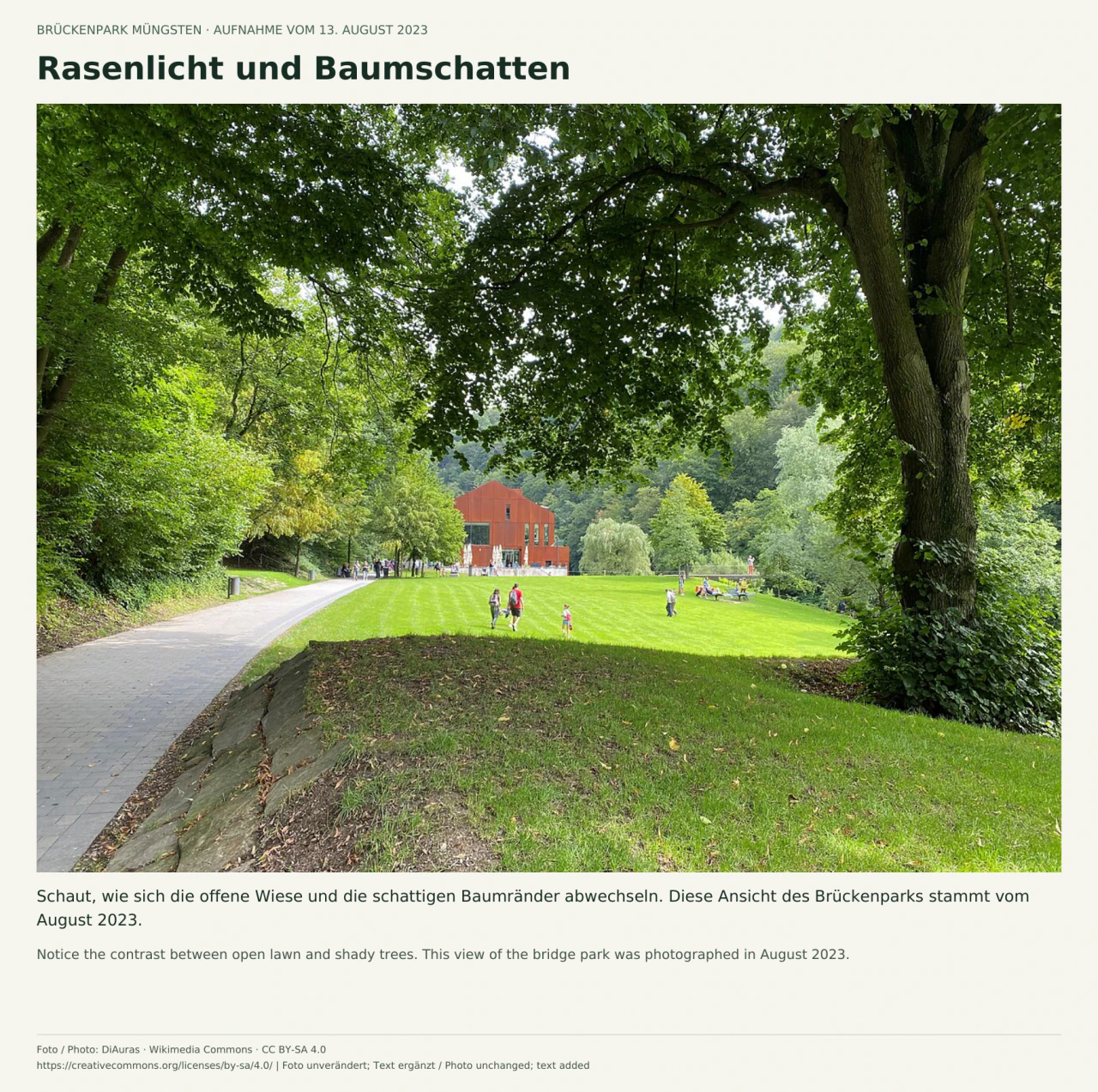 Unter einem großen Baum führt links ein gepflasterter Weg an einer sonnigen Rasenfläche vorbei; kleine Besuchergruppen und ein rostrot verkleidetes Gebäude stehen im Hintergrund.