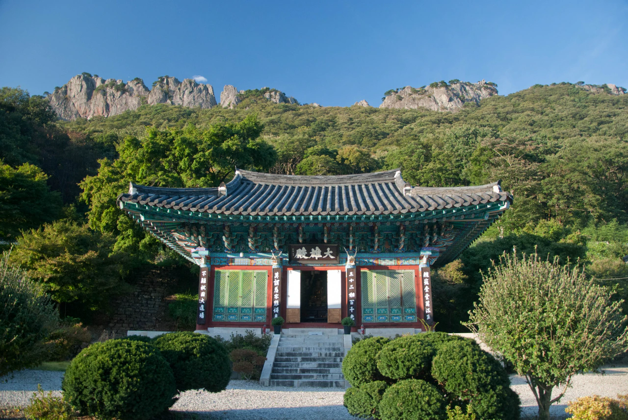 A painted traditional temple building with a grey tiled roof and stone steps stands below green woodland and a serrated pale rock ridge.