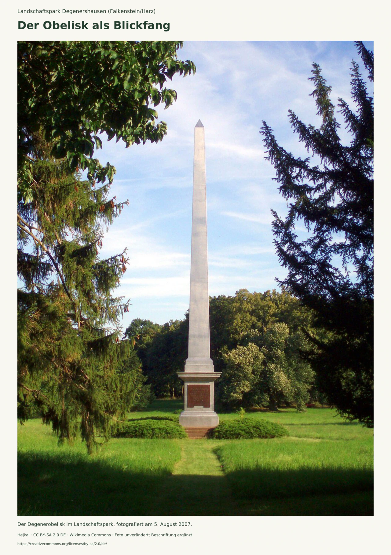 Ein schmaler heller Obelisk mit spitzer Spitze und dunkler Tafel am Sockel steht in einer Rasenfläche, seitlich von hohen Bäumen eingerahmt.