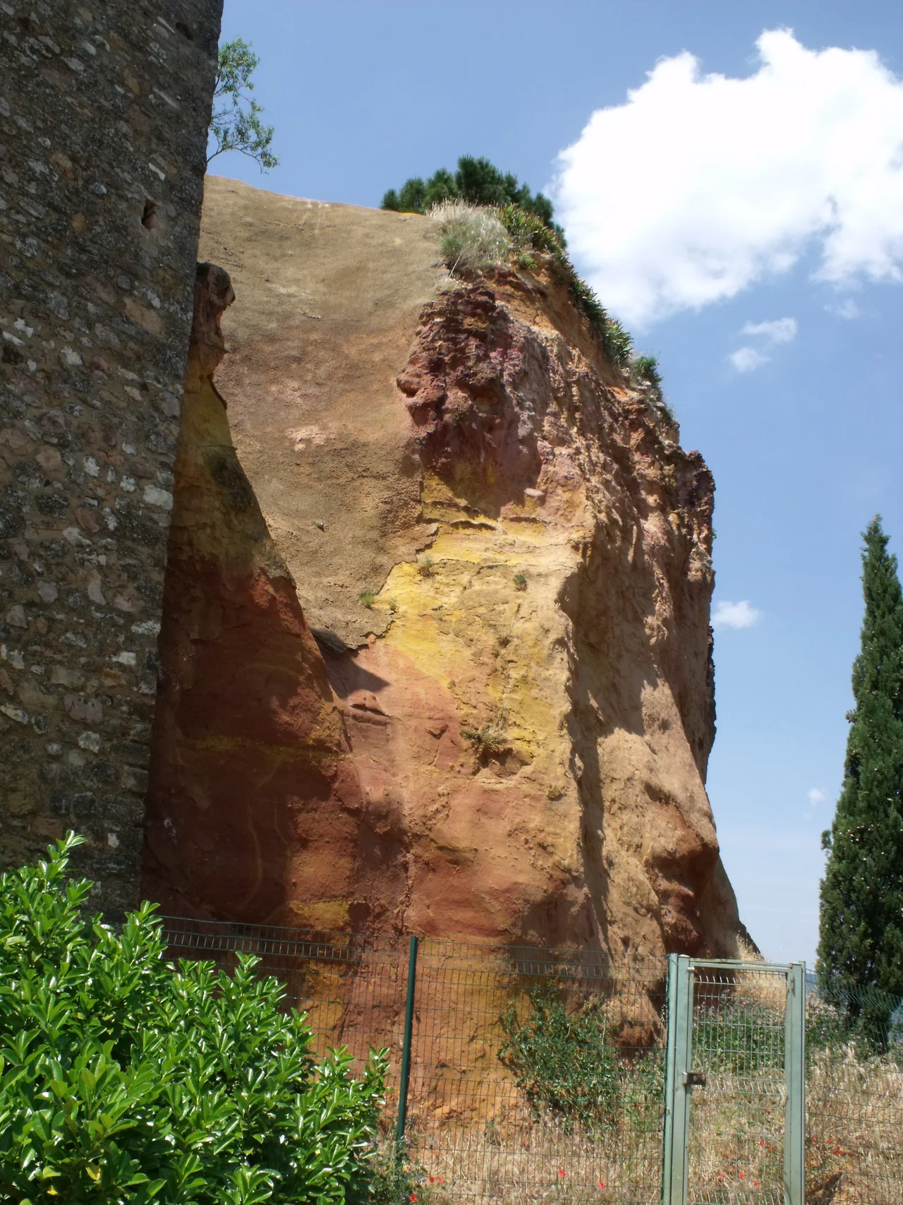 A tall yellow, orange and red ochre face rises beside a stone building, with a wire fence, gate and shrubs below and blue sky above.