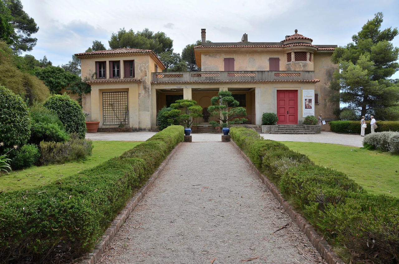 A hedge-lined gravel path leads to an ochre villa with red shutters, surrounded by trees.