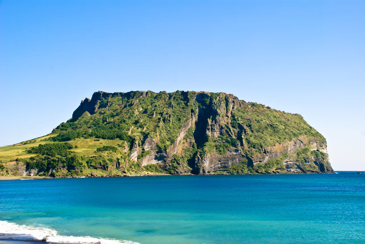 A steep, green-covered volcanic cone with exposed layered cliffs beside blue sea under a clear sky.