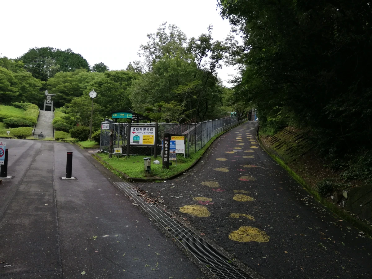 A paved uphill path with painted footprints runs beside trees and a fenced slope-car station; another path and steps lie to the left.