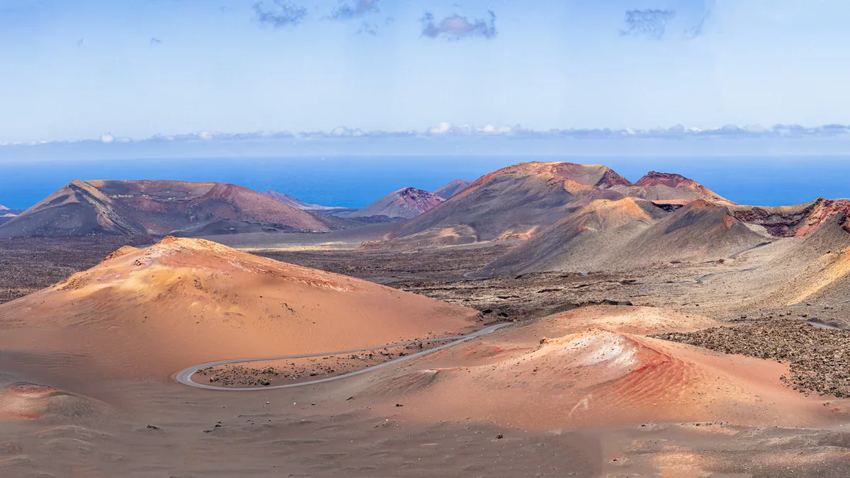 Red, orange and dark volcanic hills surround a winding paved road, with the blue sea beyond.