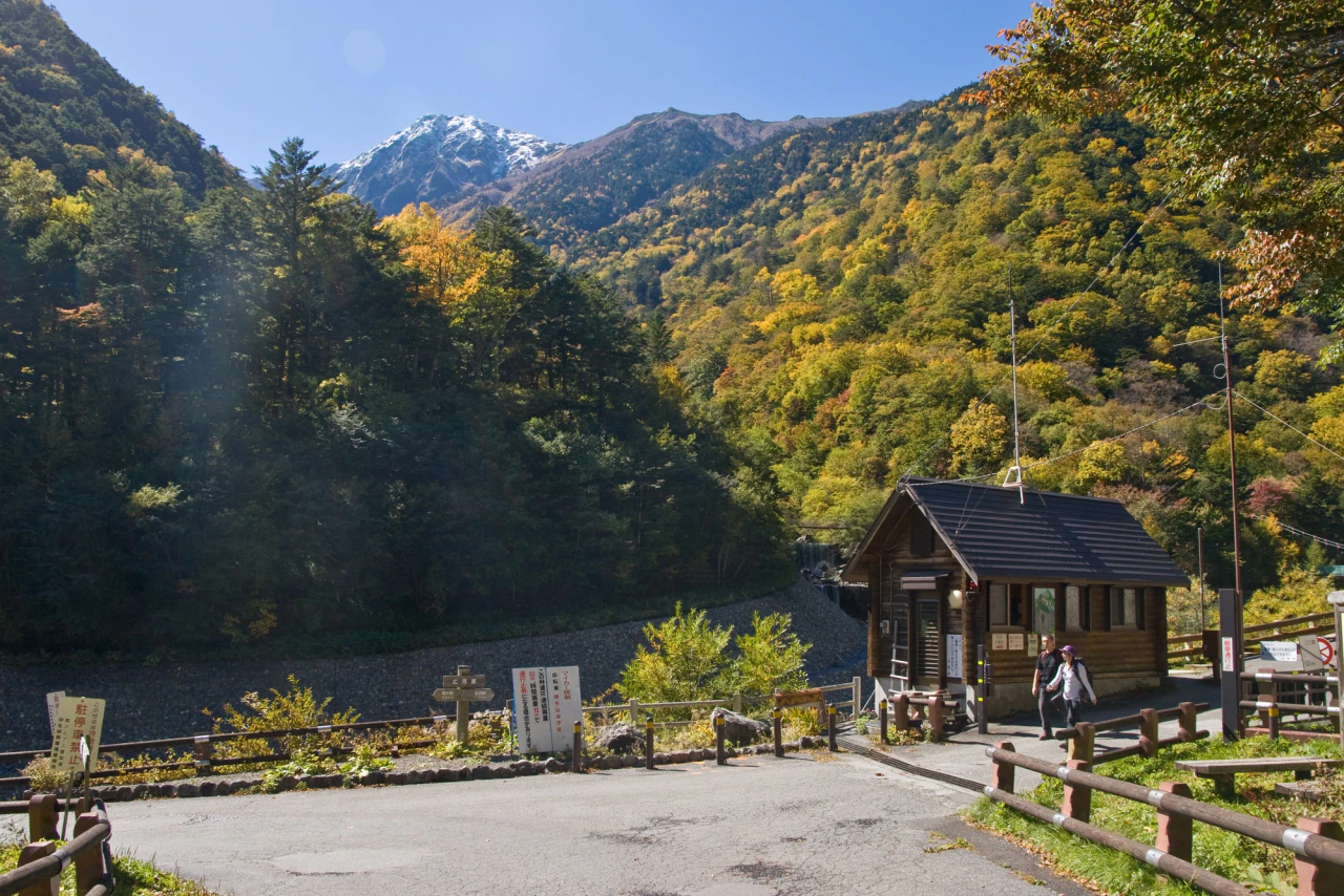 A snow-dusted mountain rises above autumn-coloured wooded slopes, with a small wooden building, roadside railings and visitors in the foreground.