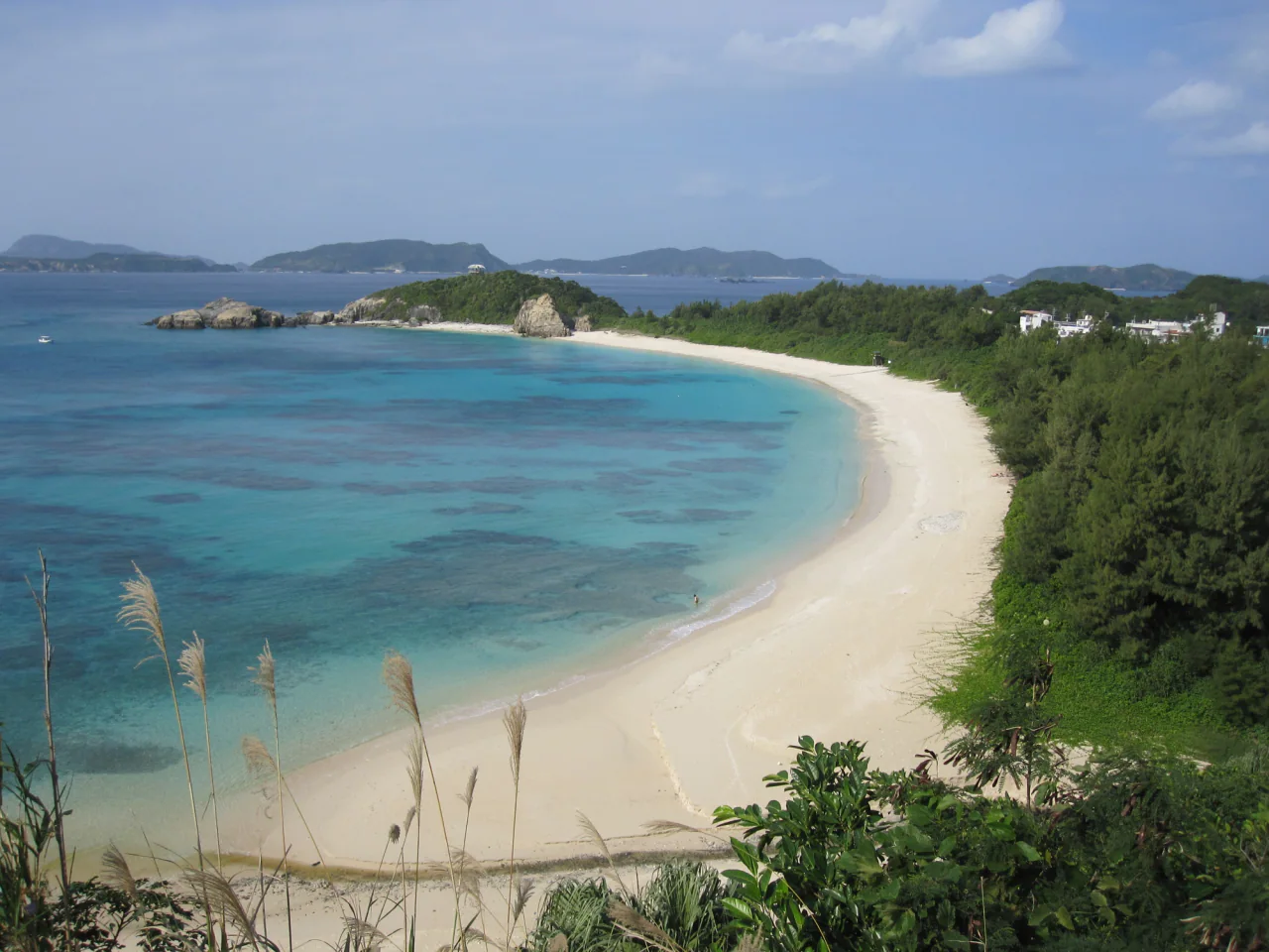 A pale sandy beach curves around turquoise water, backed by dense greenery and scattered buildings, with islands across the bay.
