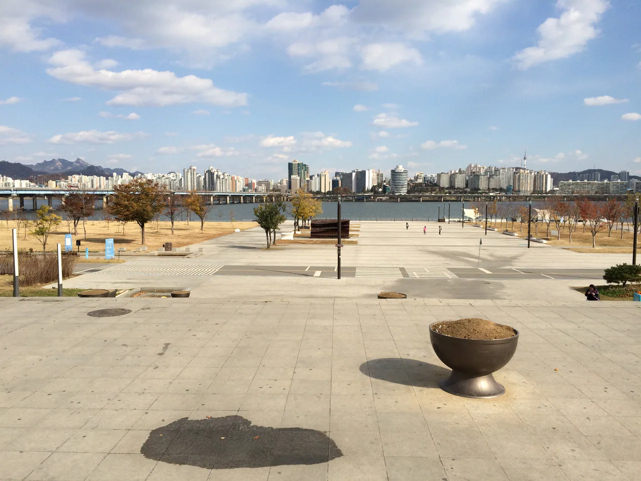 Broad paved steps and a plaza bordered by lawns and trees, with the Han River, a bridge and distant buildings beyond.