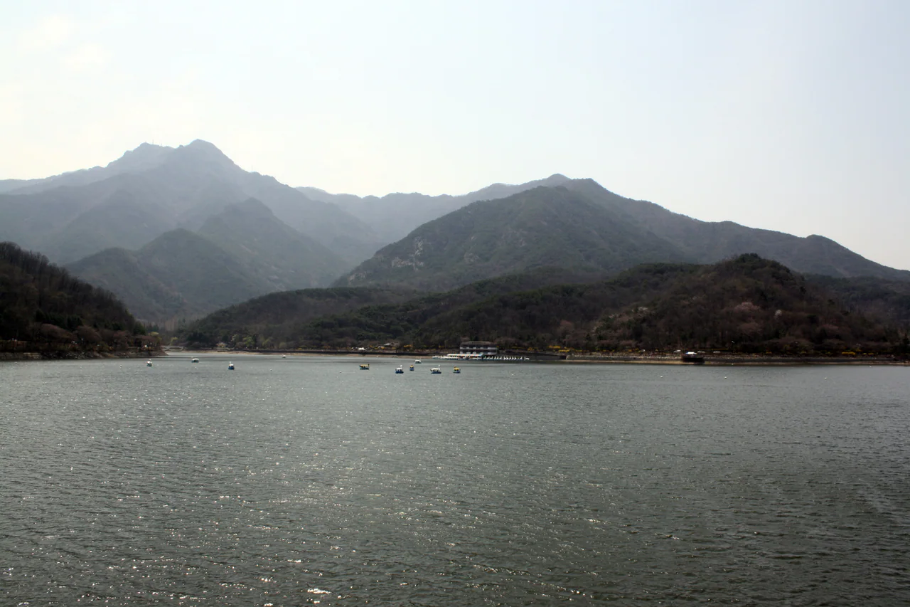 Small boats dot a broad reservoir beneath wooded mountain ridges and a pale, hazy sky.