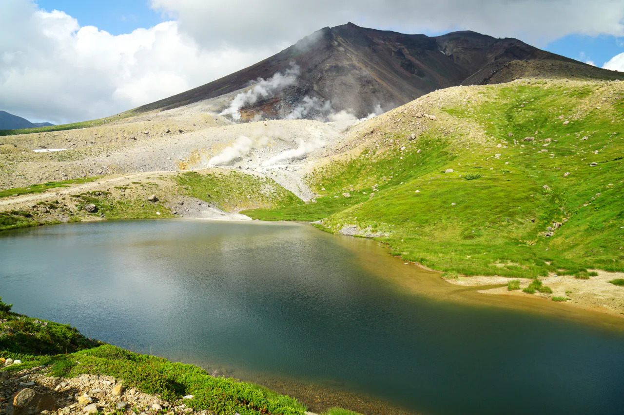 A dark blue-green pond lies below green and pale rocky slopes, with white fumarole plumes rising beneath Asahidake’s rocky summit.