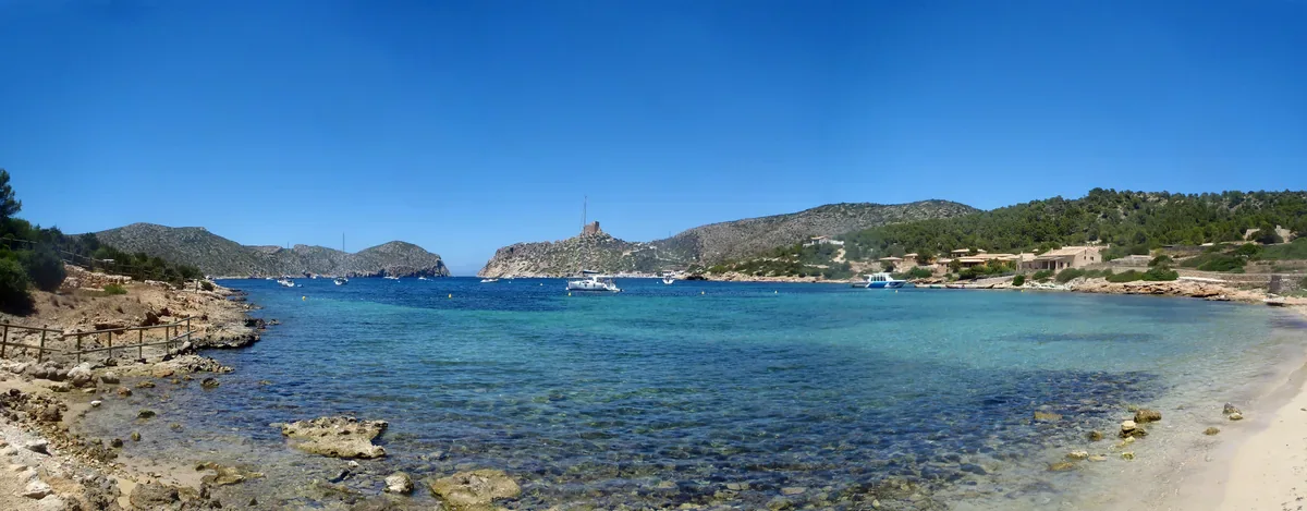 Clear blue harbour water bordered by rocky slopes, low vegetation and shore buildings.
