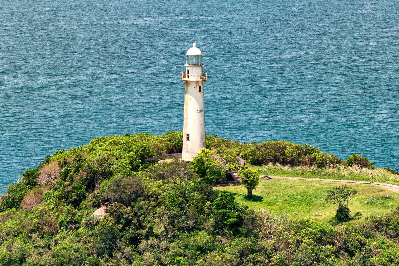 A pale lighthouse on a green coastal headland, with the blue sea behind it and a small visitor on a path to the right.