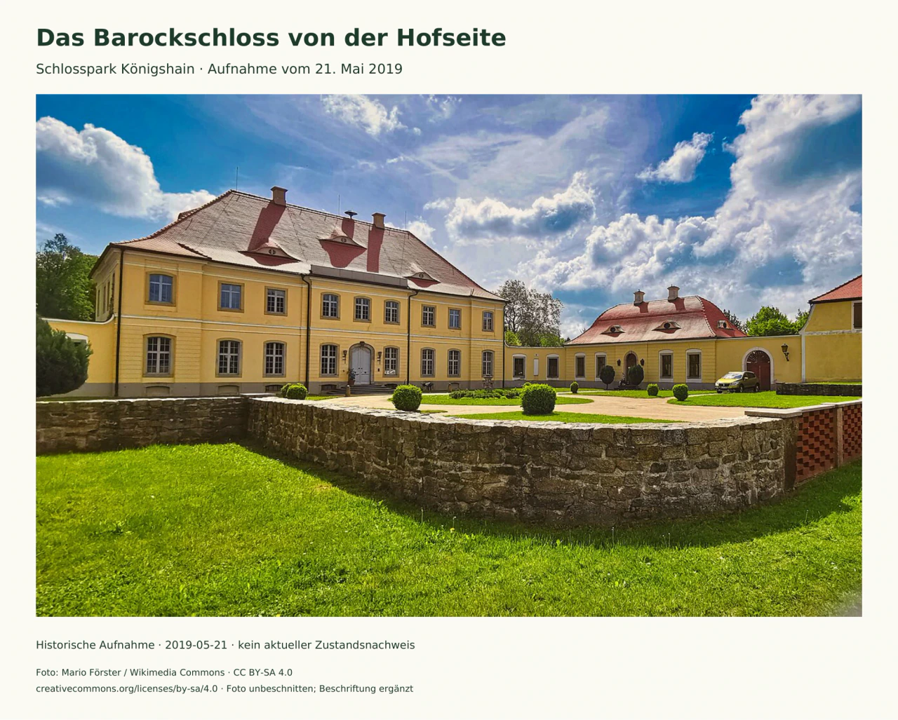 Gelbes Barockschloss mit rotem Dach und Seitenflügeln; davor ein Hof mit rund geschnittenen Sträuchern, eine niedrige Steinmauer und Rasen.