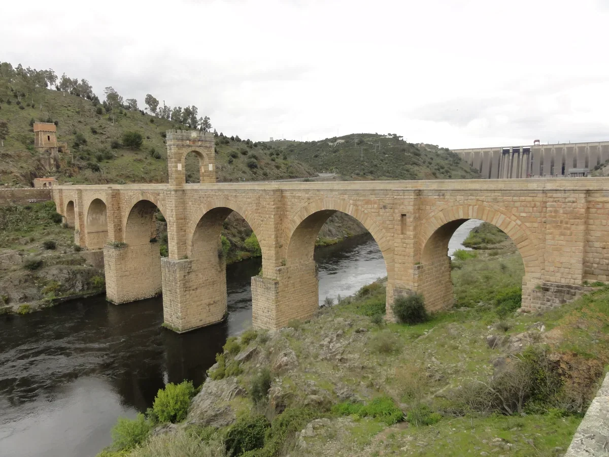 The stone arches and central monumental arch of Alcántara’s Roman bridge over the Tagus, with the modern dam behind.