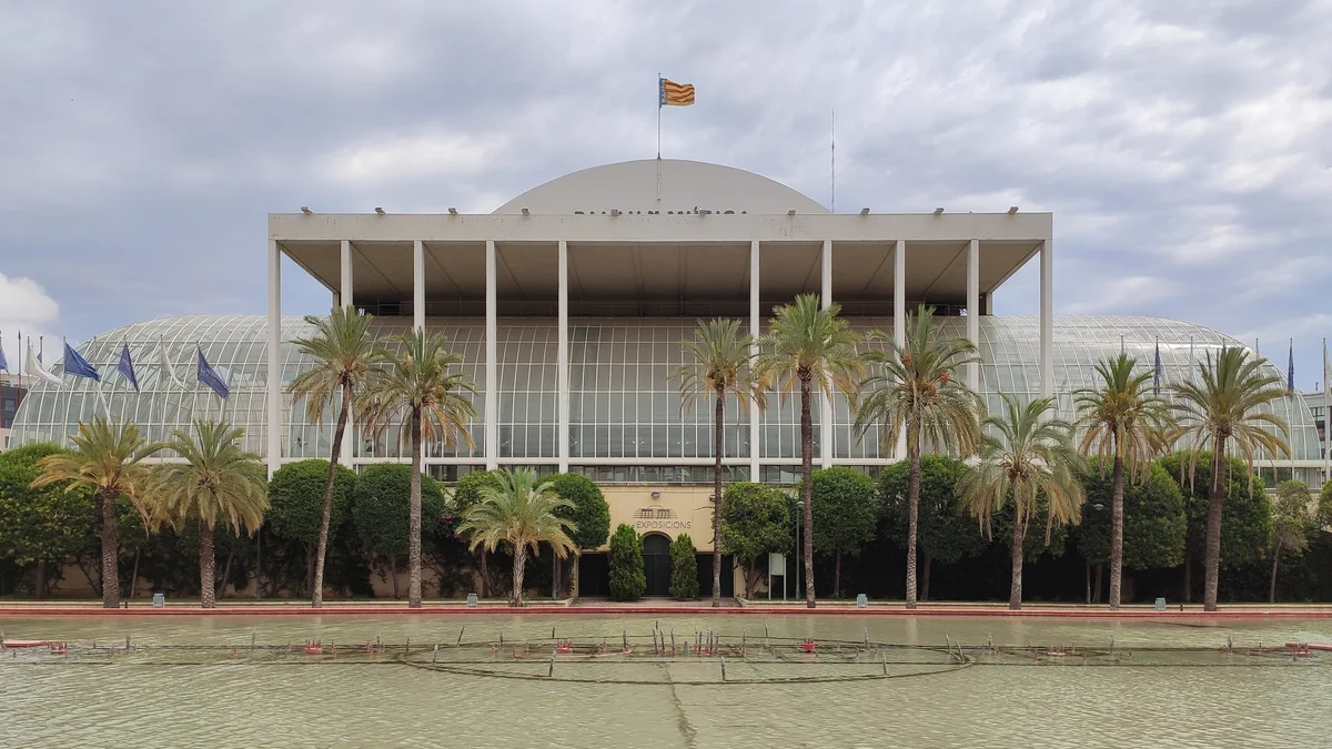 Palm trees line the glass-fronted Palau de la Música behind an ornamental pool beneath a cloudy sky.