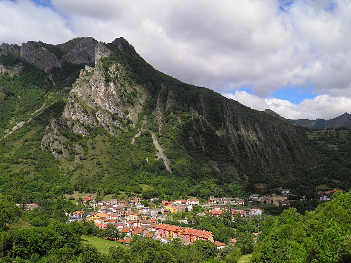 Pola de Somiedo’s village buildings sit below a steep wooded hillside and pale limestone cliffs under a cloudy sky.