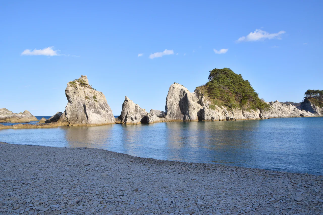 Pale jagged rock formations rise from blue water beside a white pebble beach, with green trees on the larger outcrop and a clear blue sky above.