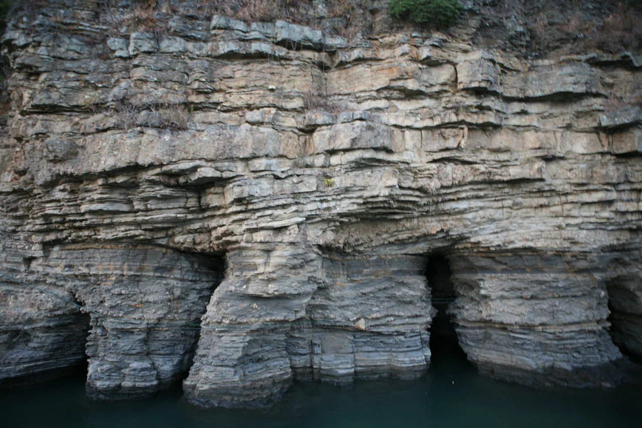Thin grey rock layers form a coastal cliff, with dark recesses at its base above blue-green seawater.