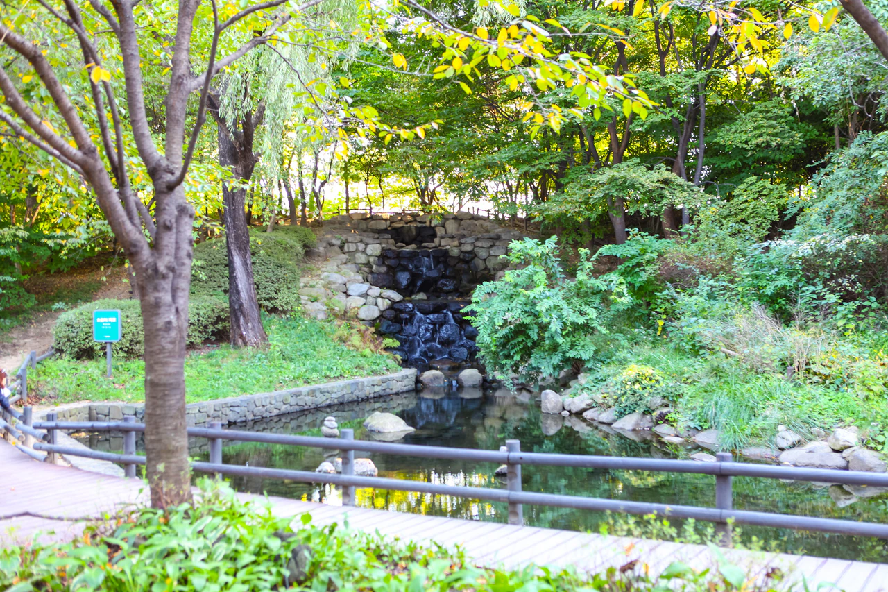 Trees surround a small stone cascade and pool, with a wooden railing across the foreground and a green sign to the left.