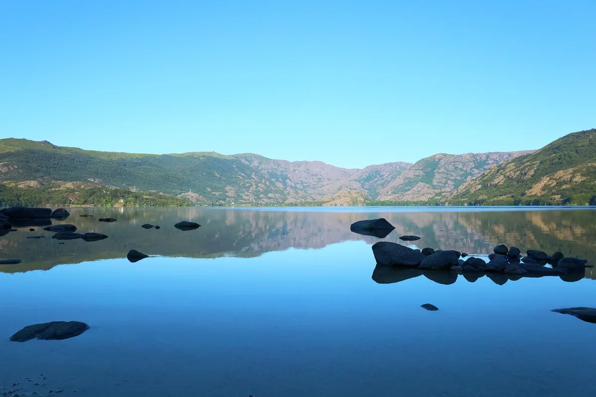 Dark shoreline rocks stand in a calm blue lake, with green and rocky hills reflected beneath a clear sky.