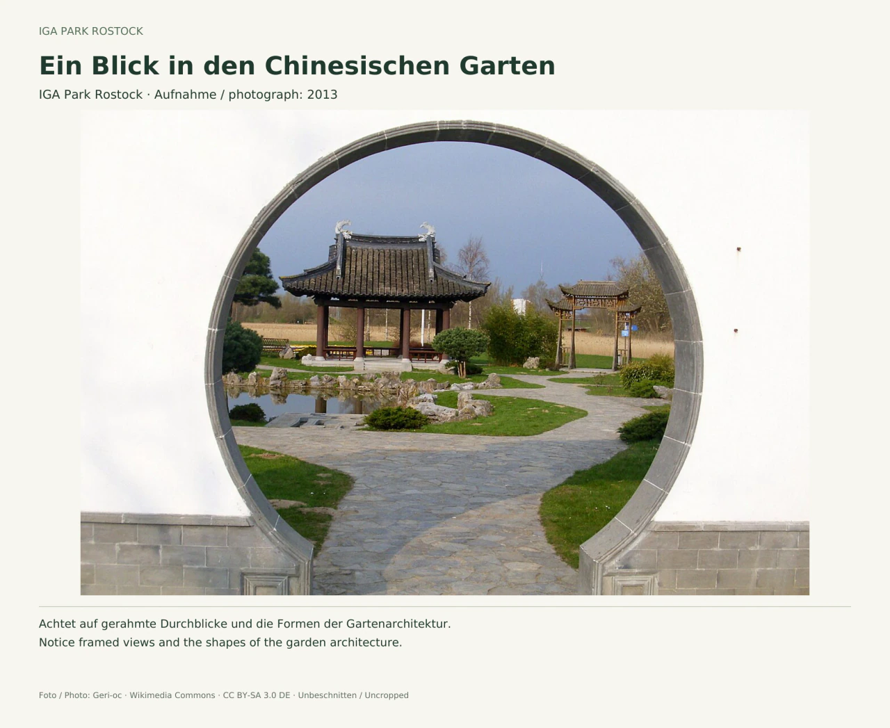 Blick durch eine große runde Öffnung in einer weißen Gartenmauer: Dahinter liegen ein gepflasterter Weg, ein kleiner Teich, Felsen, Sträucher und ein Pavillon mit dunklem geschwungenem Dach.