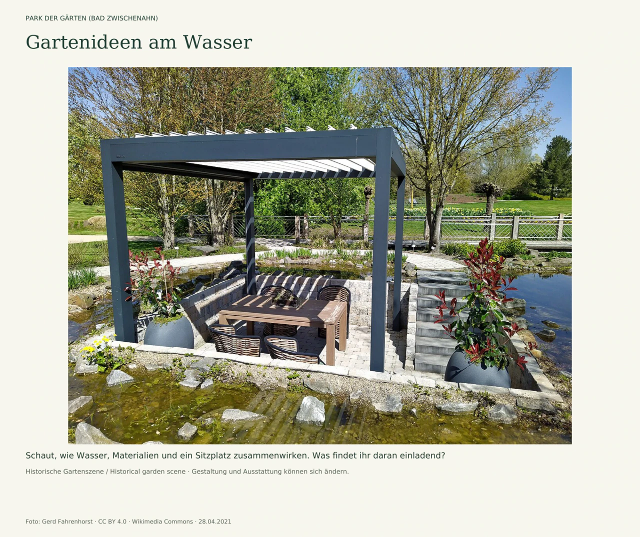 Graue Pergola über einem abgesenkten Sitzplatz mit Tisch und vier Stühlen. Rechts führen Stufen hinab; flaches Wasser, Steine und Pflanzkübel umgeben den Platz. Dahinter verläuft eine Brücke zwischen Bäumen.