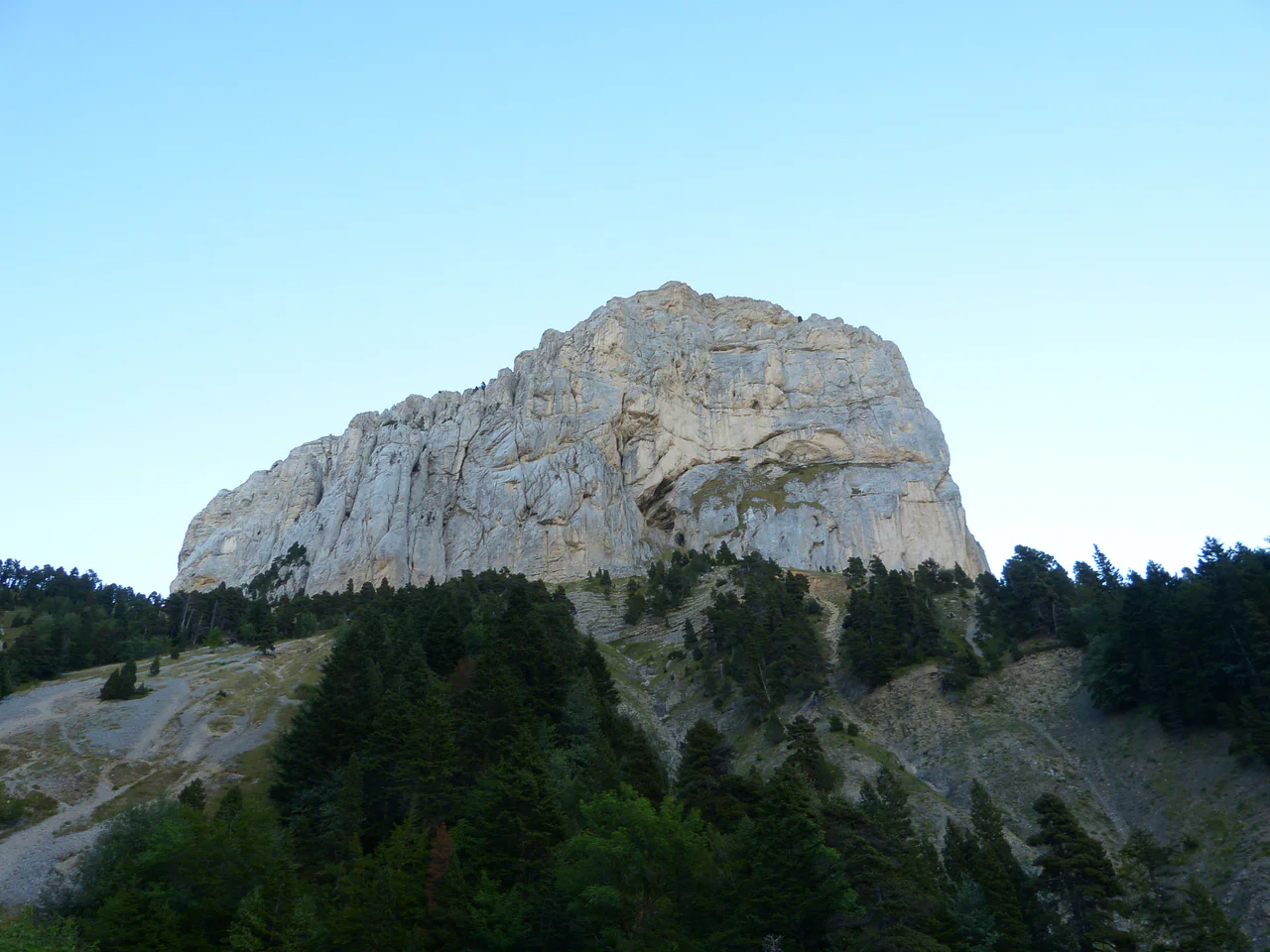 A massive pale limestone wall rises above dark woodland and sloping scree beneath a clear blue sky.
