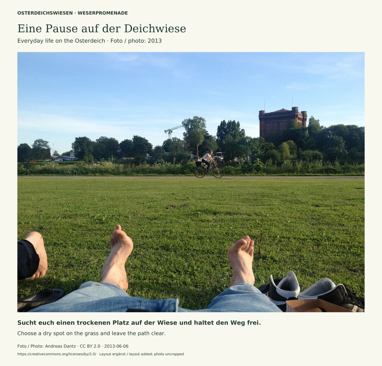 Foto aus der Perspektive einer auf der Wiese sitzenden Person: Füße und Schuhe im Vordergrund, dahinter grüner Rasen, ein vorbeifahrender Radfahrer, Uferpflanzen und der rote Backstein-Wasserturm auf dem Stadtwerder.
