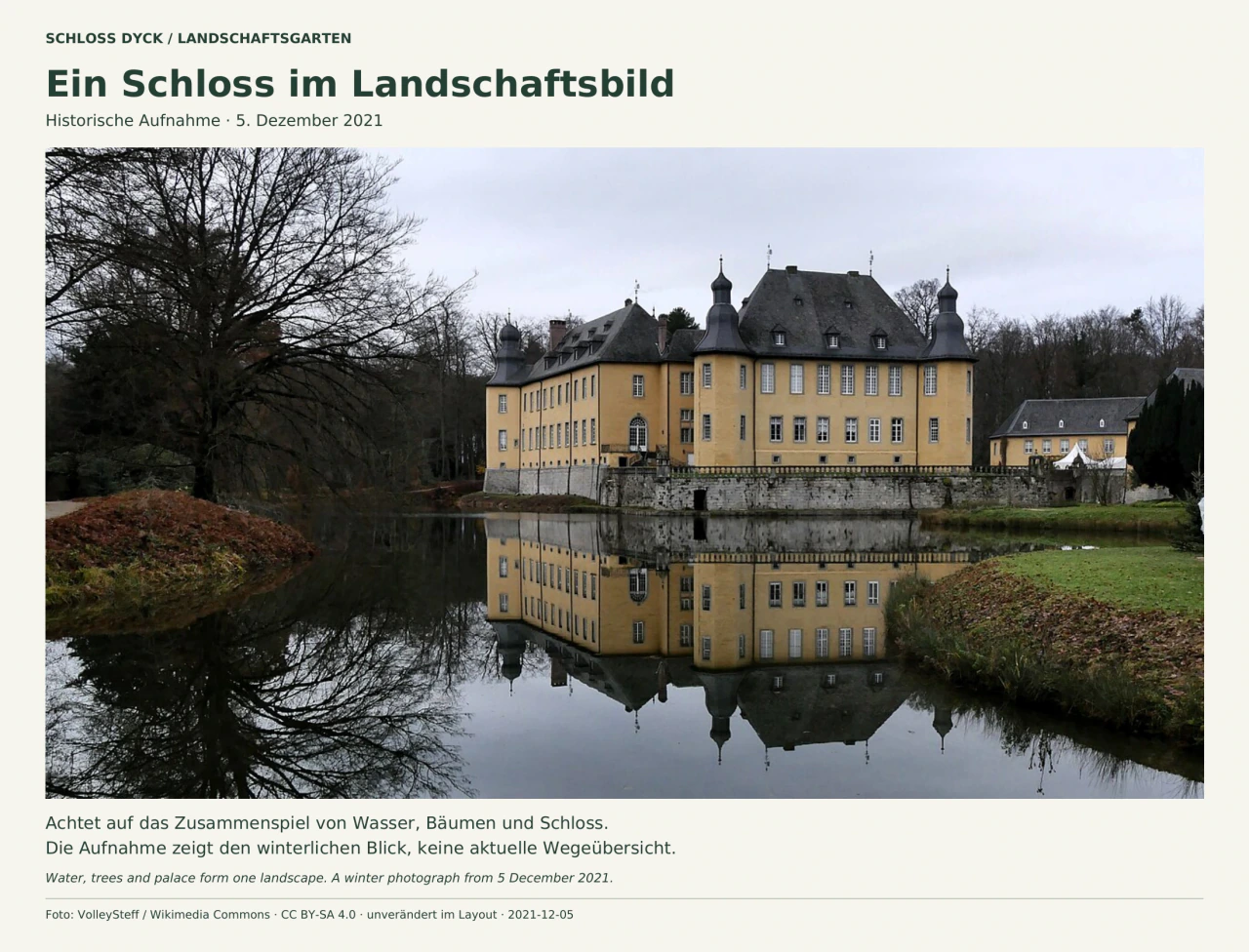 Gelbes Schloss Dyck mit dunklen Dächern und mehreren Türmchen hinter einem ruhigen Wassergraben. Schloss und kahle Bäume spiegeln sich im Wasser; braunes Laub bedeckt Teile der geschwungenen Ufer. Die Beschriftung datiert die Aufnahme auf Dezember 2021.