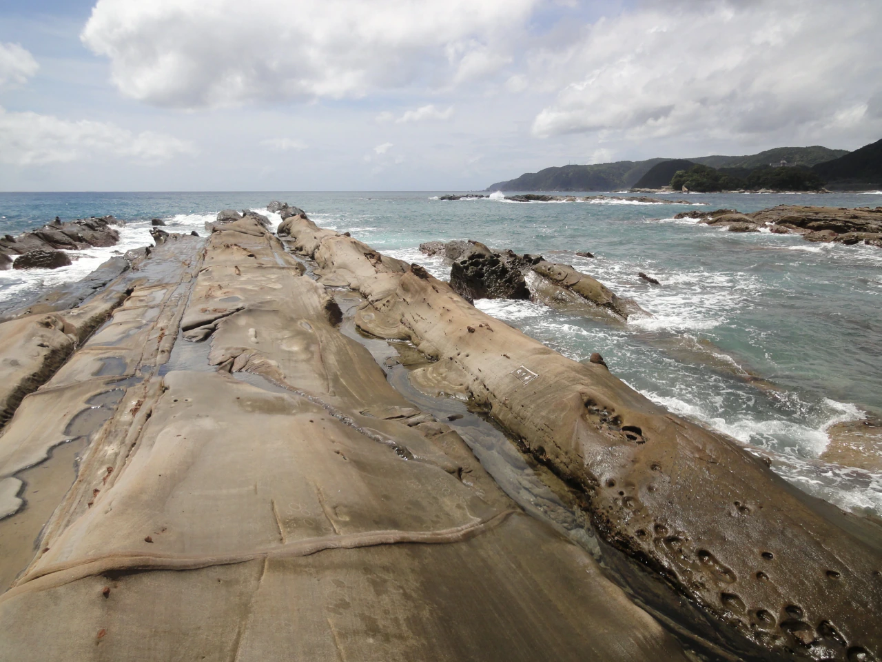 Broad tan rock ribs with seams and small hollows extend toward breaking waves, with a green headland across the water.