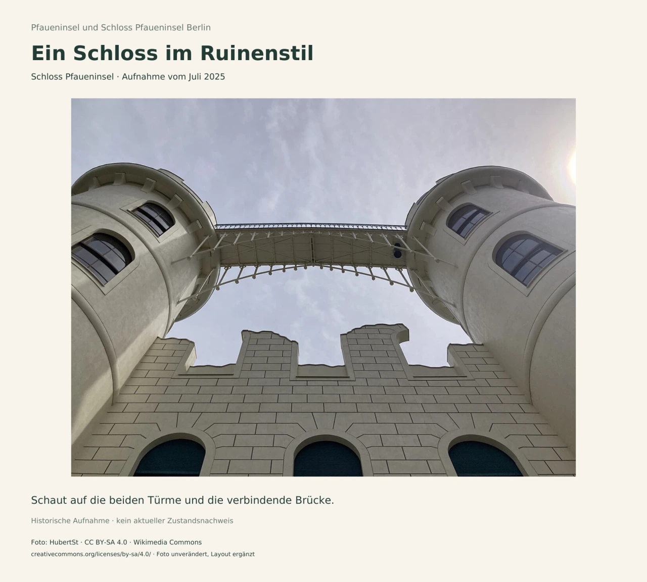Blick steil nach oben zwischen zwei hellen runden Schlosstürmen zur verbindenden Metallbrücke; darunter eine Wand mit unregelmäßiger Oberkante.