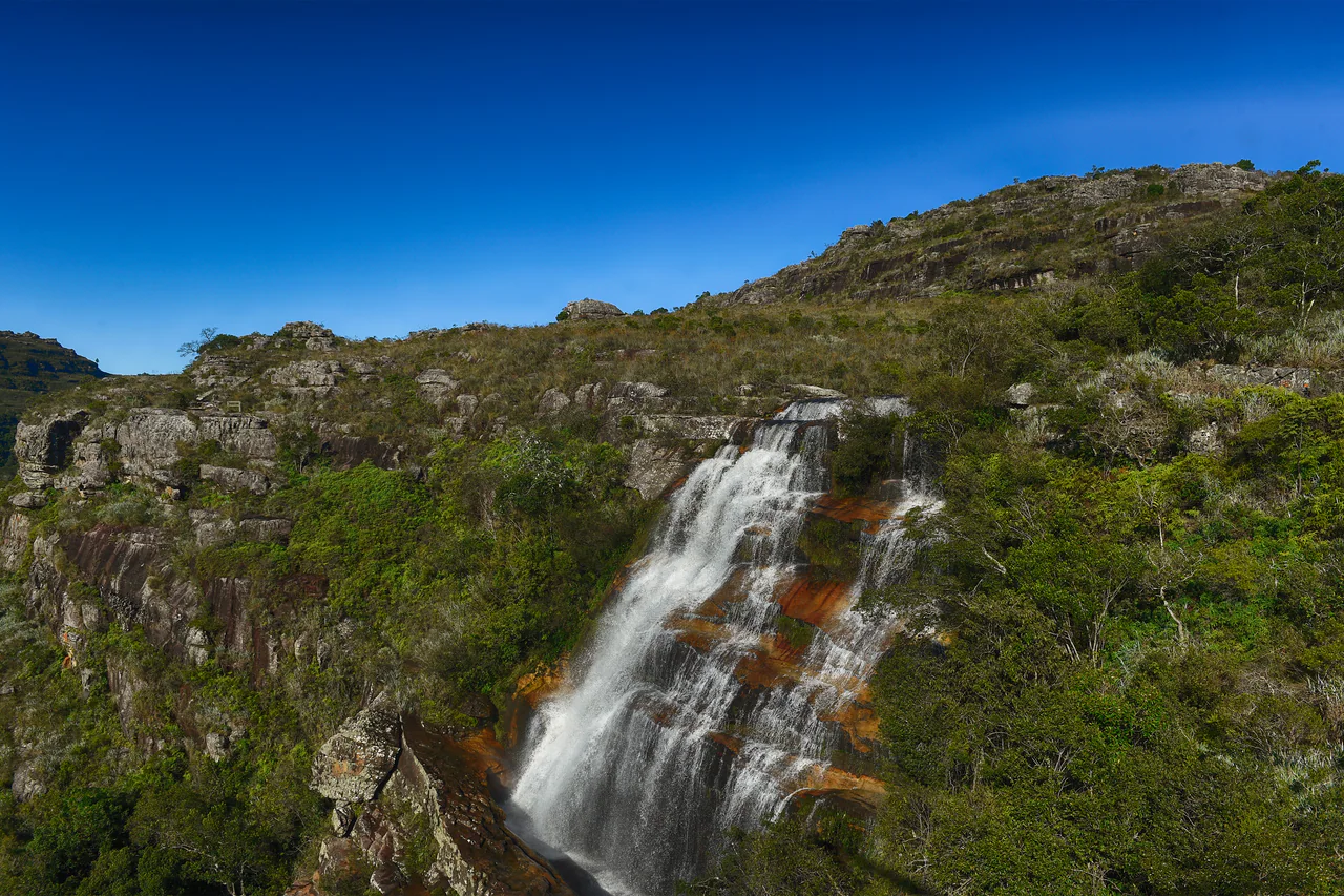 White water cascades down layered orange-brown rock amid green vegetation, with a rocky grass-covered ridge and clear blue sky above.