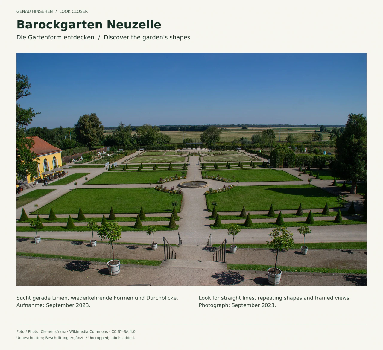 Blick von einer Terrasse über den Barockgarten Neuzelle im September 2023: symmetrische Rasenflächen und gerade Wege um ein rundes Becken, geschnittene Gehölze und Kübelpflanzen; dahinter liegt die offene Landschaft.