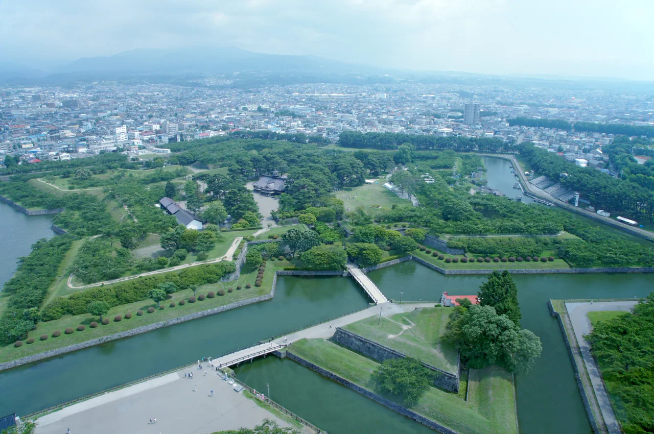 An elevated view of angular moats, stone-faced earthworks, green trees and the central reconstructed Magistrate’s Office, with Hakodate beyond.