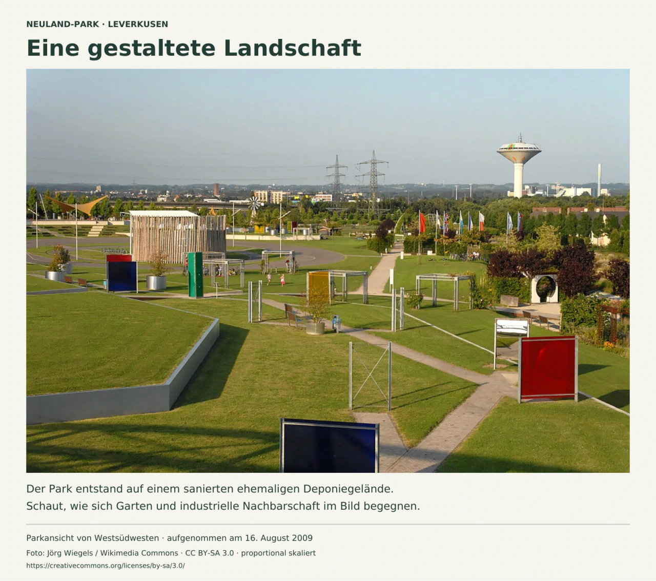 Blick über geometrische Rasenflächen und Wege des Neuland-Parks mit farbigen Gartenelementen. Hinter den Bäumen stehen Strommasten, Gebäude und ein hoher Wasserturm; die Aufnahme stammt von 2009.