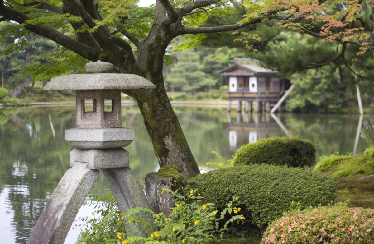 A two-legged stone lantern stands beneath a leafy tree beside a pond, with a waterside pavilion in the background.