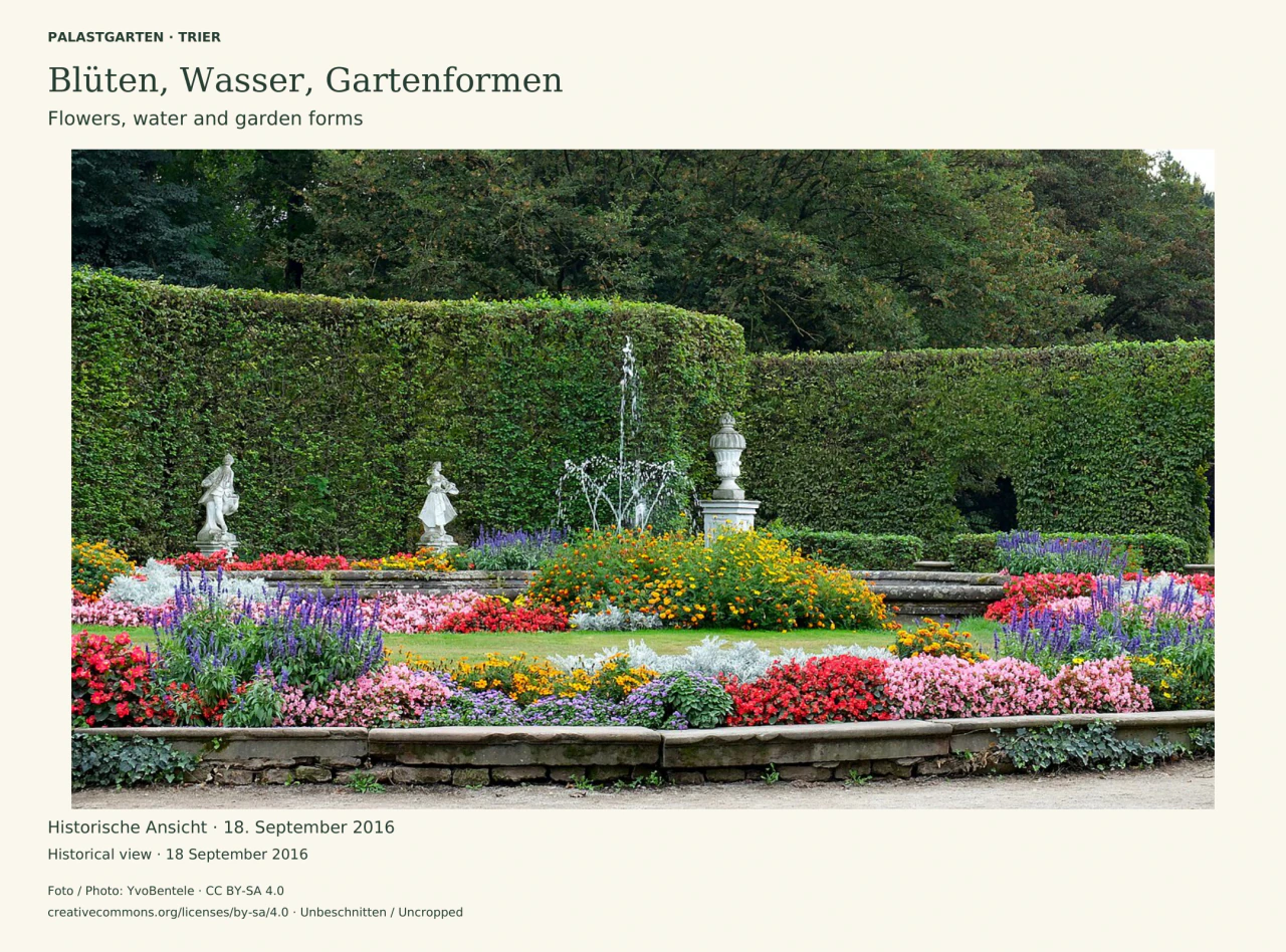 Bunte Blumenbeete in Rosa, Rot, Gelb und Violett umgeben einen Rasenabschnitt und ein flaches Brunnenbecken mit aufsteigenden Wasserstrahlen. Dahinter stehen helle Skulpturen und eine Vase vor hohen geschnittenen Hecken.