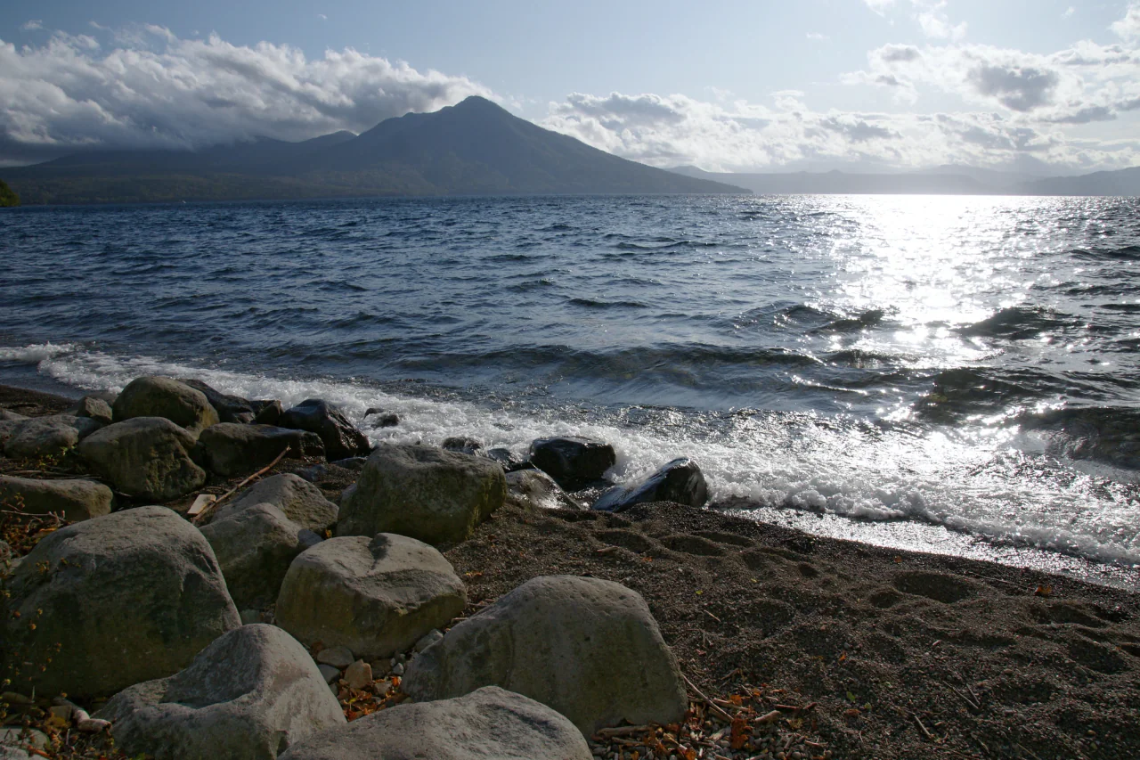 Waves break against dark shore pebbles and large rocks, with a forested mountain across Lake Shikotsu beneath scattered clouds and bright sunlight.