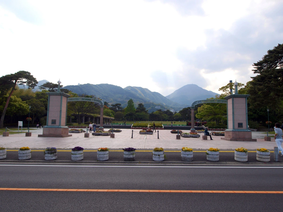 Two tall entrance pillars frame an open plaza with flower planters, trees and a mountain backdrop; a road crosses the foreground.