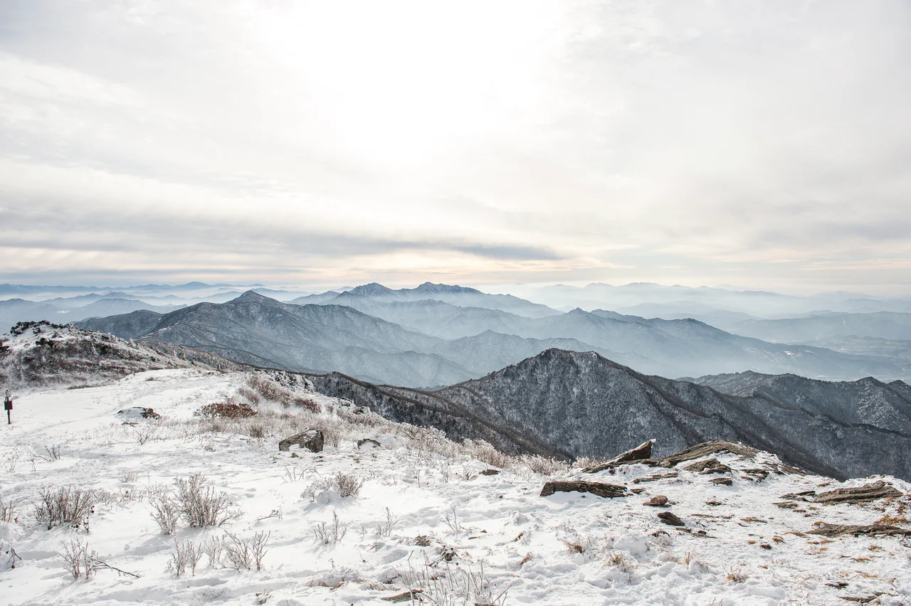 Snow covers an open rocky foreground and low shrubs, with successive dark mountain ridges fading into pale haze beneath a broad cloudy sky.