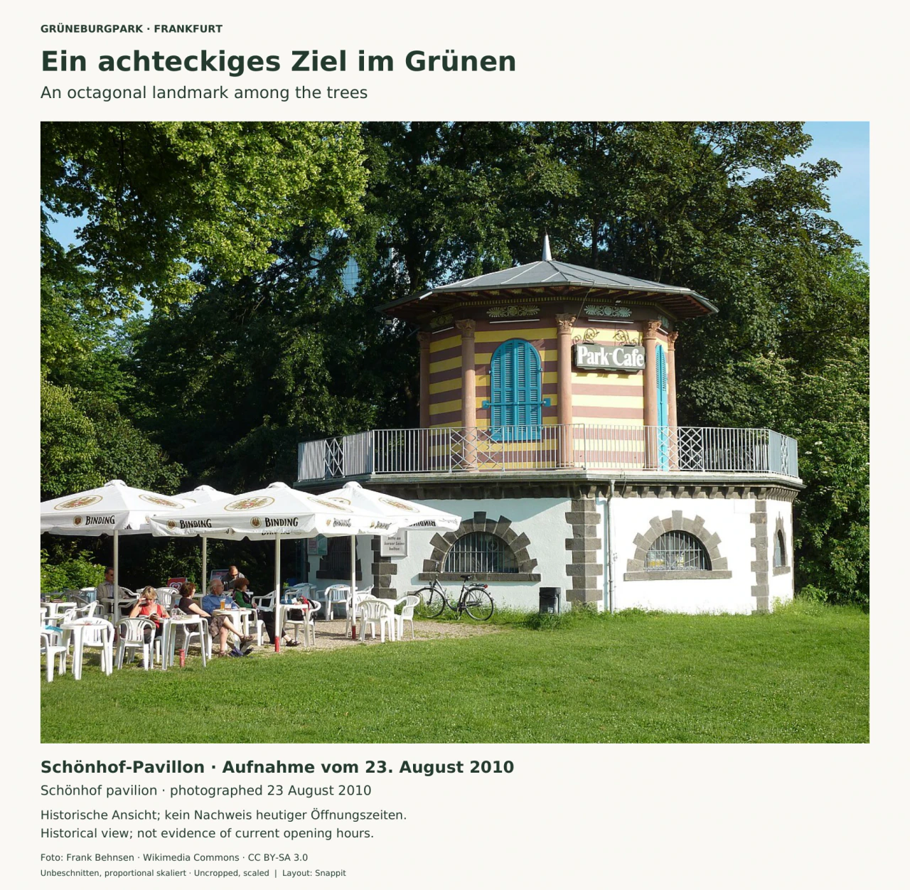 Ein achteckiger Pavillon mit gelb-rosa gestreiften Wänden, blauen Fensterläden und grauem Dach zwischen hohen Bäumen; davor sitzen Menschen unter weißen Sonnenschirmen.