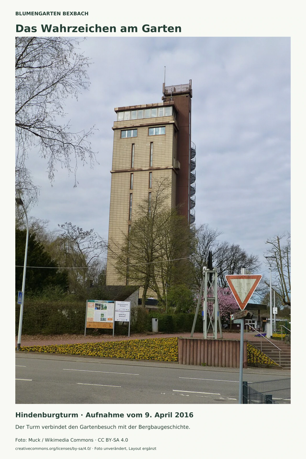 Der hohe rechteckige Hindenburgturm ragt hinter Bäumen und einem gelben Blumenstreifen auf. Oben liegt eine verglaste Etage; rechts ist eine äußere Wendeltreppe zu sehen. Im Vordergrund verlaufen eine Straße und ein Vorfahrt-gewähren-Schild.