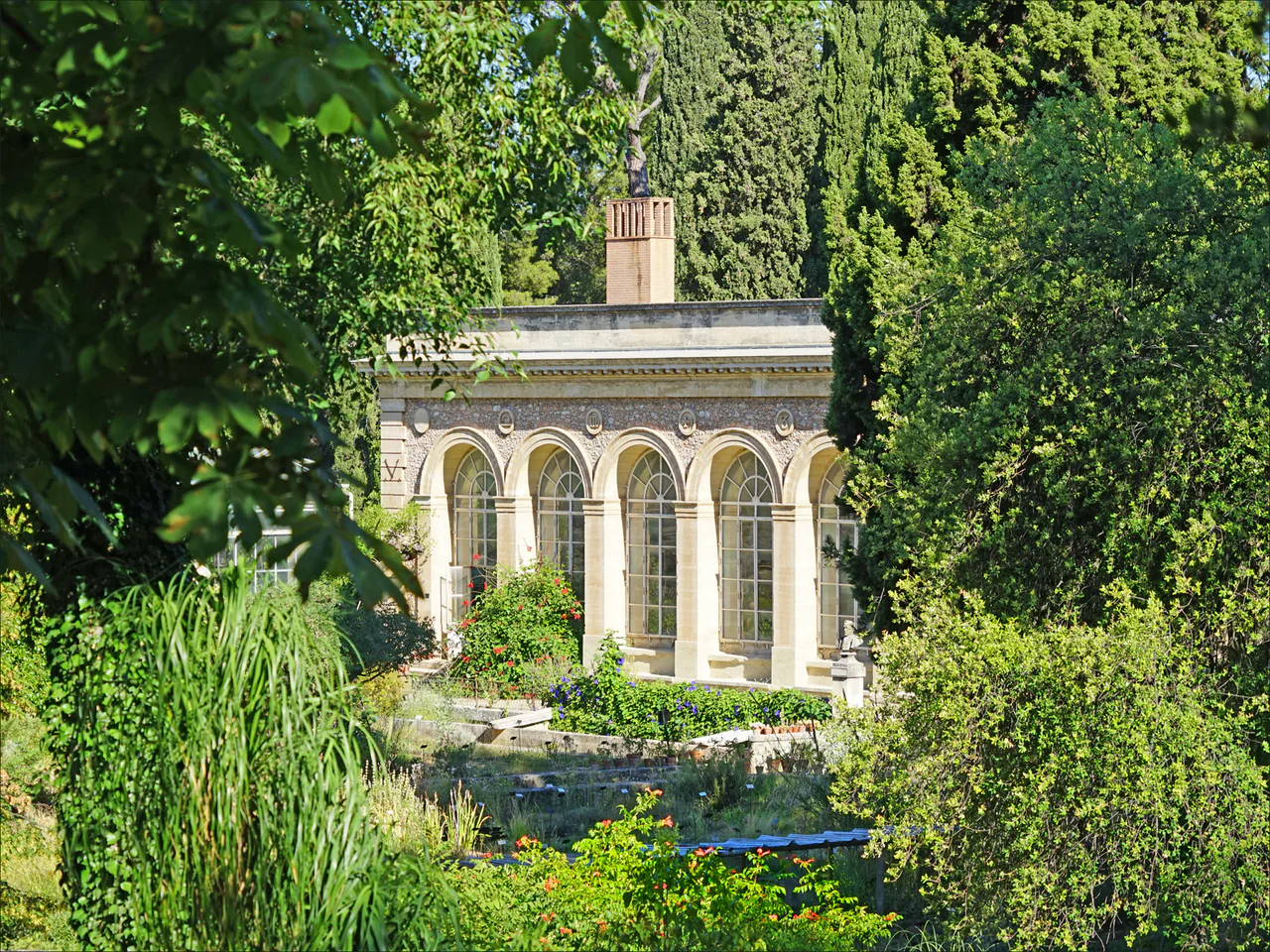 An orangery with tall arched windows appears between dense trees, shrubs and planted garden beds.