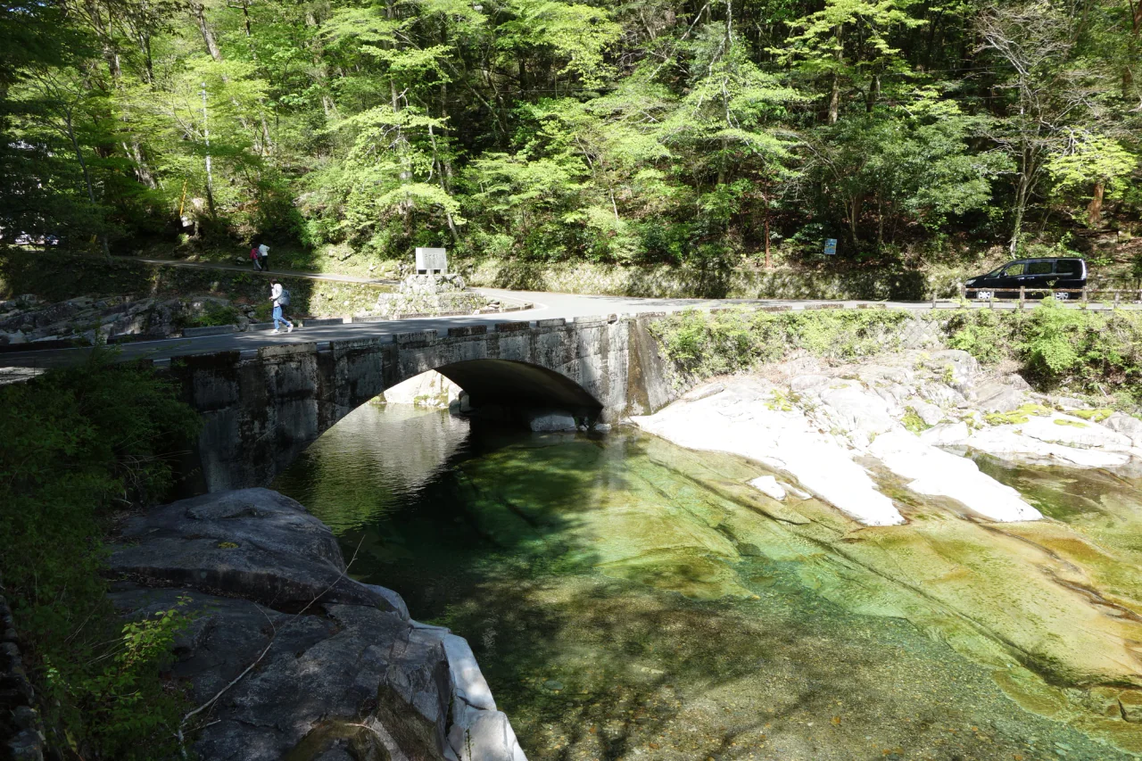 A low arched bridge crosses green-tinted water between pale rock slabs and leafy woodland.