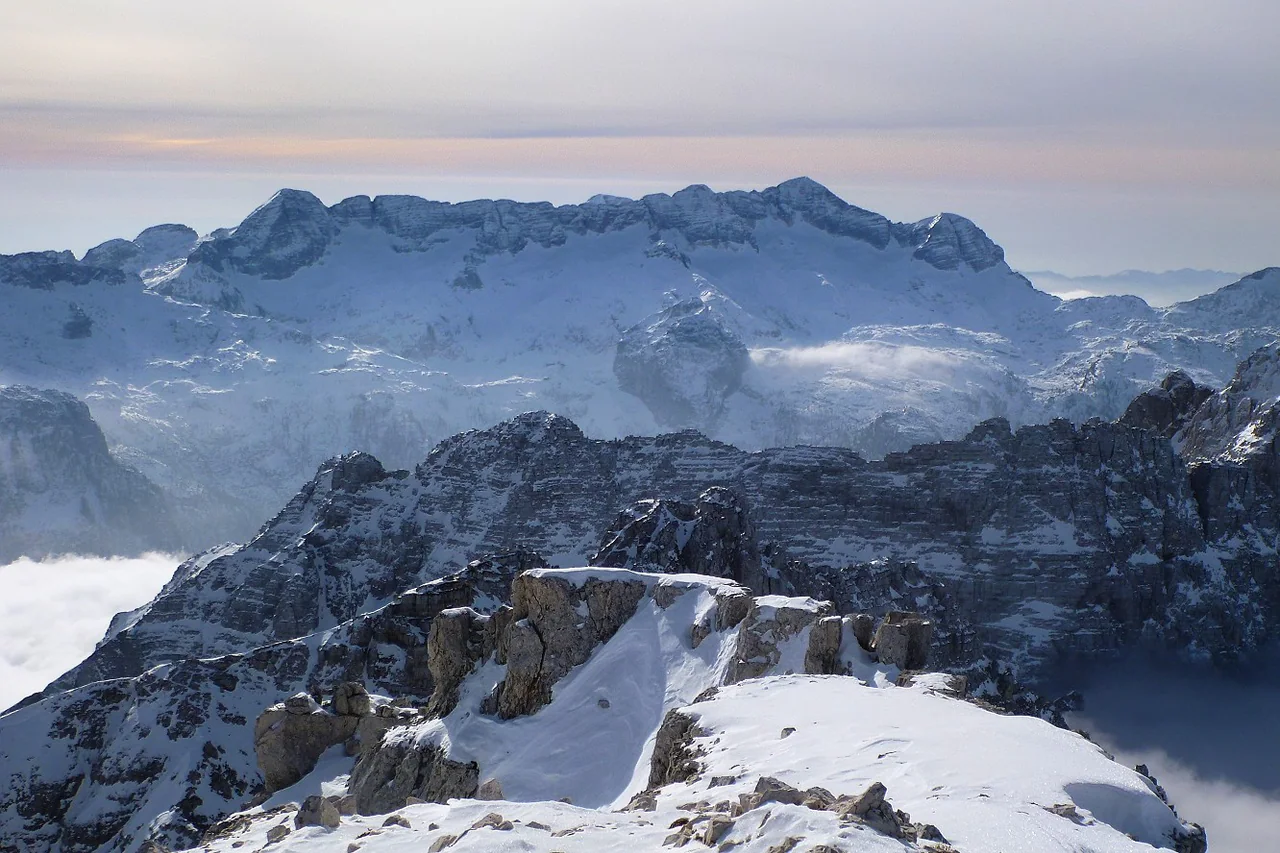 Snow covers a succession of jagged limestone ridges beneath a pale sky, with low cloud between the mountains.