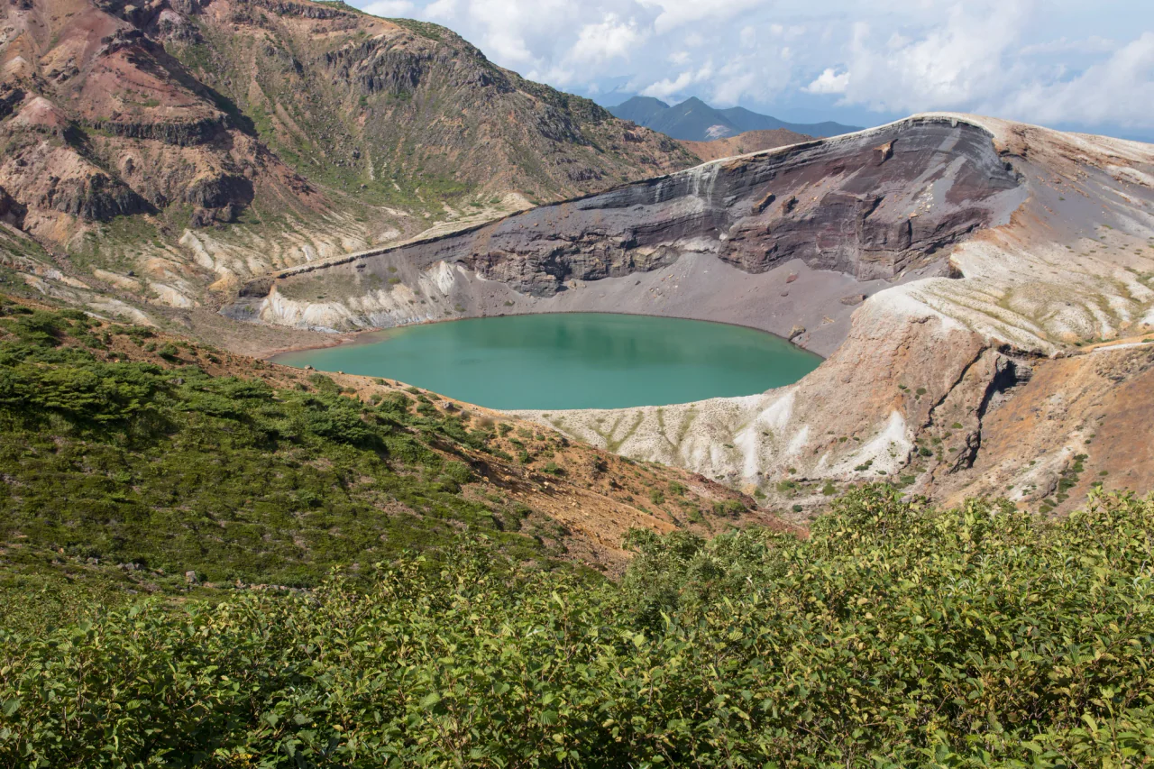 A green-blue crater lake lies between steep, layered volcanic walls, with shrubs in the foreground.