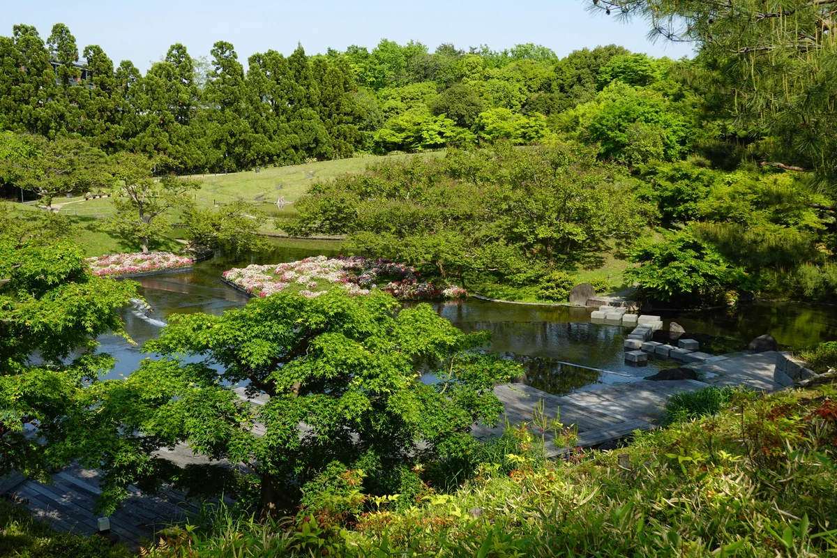 Spring-green trees surround dark shallow water, pink and white flowering beds, stone steps and a grassy slope in Umekoji Park’s Suzaku-no-niwa garden.