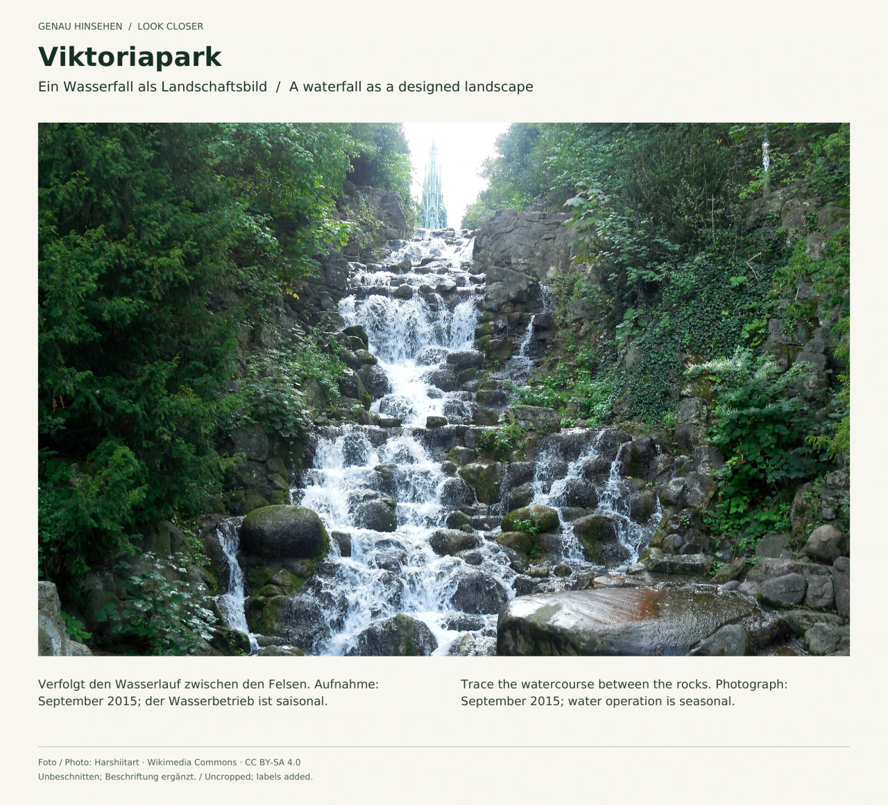 Blick den künstlichen Wasserfall im Viktoriapark hinauf: Wasser fließt über unregelmäßige Felsstufen zwischen dichtem Grün; oben ist die Spitze des Nationaldenkmals zu sehen. Aufnahme von September 2015.