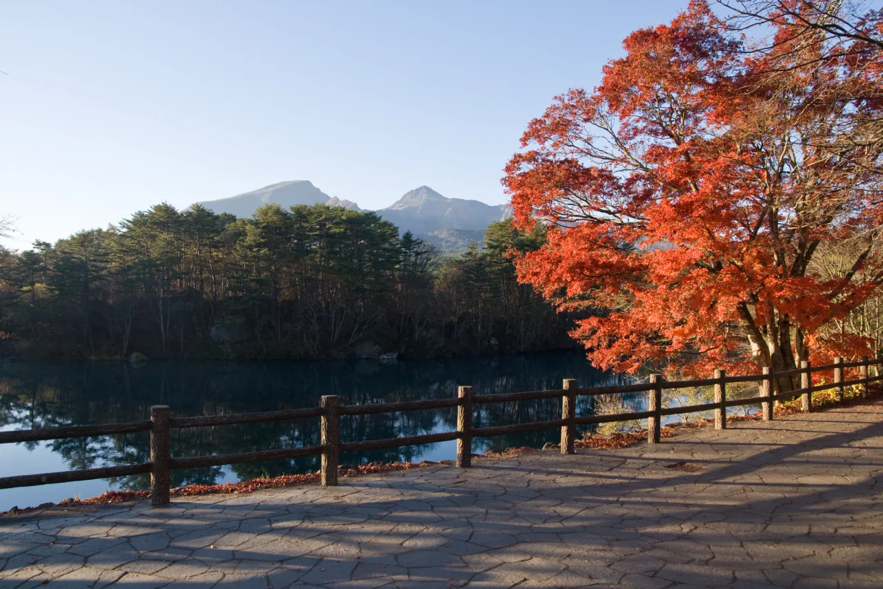 Red-orange leaves overhang a fenced stone path beside a dark reflective pond, with forest and the craggy profile of Mount Bandai beyond.