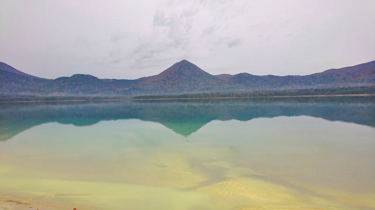 Still green-blue lake water reflects a wooded mountain ridge beneath a pale cloudy sky, with shallow water visible in the foreground.